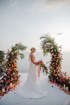 a bride standing in front of an arch with flowers and candles on the beach at sunset