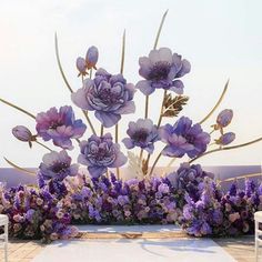 an arrangement of purple flowers and greenery in front of a white table with chairs