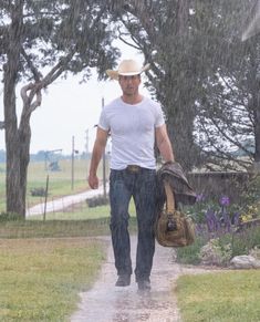 a man walking down a path in the rain with a hat and bag on his shoulder