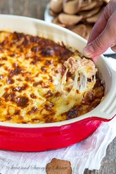 a hand holding a piece of lasagna above a casserole in a red dish