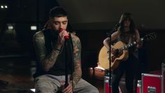 a man with tattoos sitting on a stool while holding a guitar and singing into a microphone