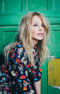 a woman with blonde hair and blue eyes posing in front of a green door wearing a floral shirt