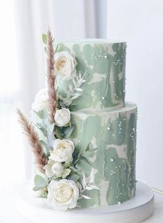 a three tiered green and white cake with flowers on the top, sitting on a table in front of a window