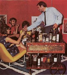 a group of people sitting around a table with liquor bottles on it and one man standing in the middle