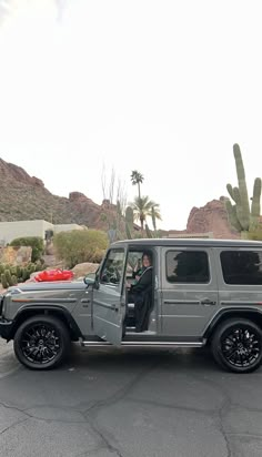 a man sitting in the driver's seat of a silver jeep parked in front of a cactus