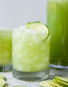 two glasses filled with cucumber and lemonade next to sliced limes on a table