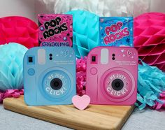 two pink and blue cameras sitting on top of a wooden board next to paper pom poms