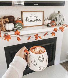 a person holding a mug in front of a fireplace with pumpkins on it and decorations