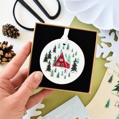 a hand holding a christmas ornament with a house in the woods on it