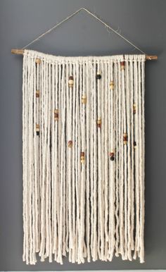 a white wall hanging with beads and wooden pegs on the bottom, in front of a gray wall