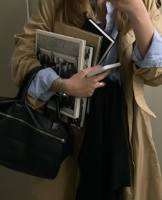 a woman in a trench coat is looking at her cell phone and holding some books