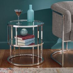 a glass side table with books and a bottle on it next to a gray chair