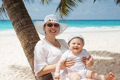 a woman holding a baby under a palm tree on the beach with blue water in the background