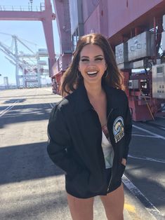 a woman is posing for the camera in front of a cargo container ship, wearing a black jacket and white panties