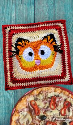 a crocheted square with an image of a cat on it next to a pizza