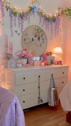 a bedroom decorated in pink and purple with flowers hanging from the ceiling, lights on the dresser