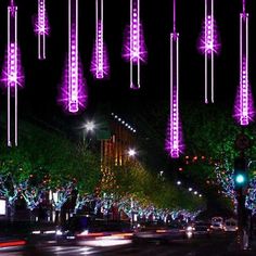 purple lights are hanging from the side of trees on a city street at night with cars passing by