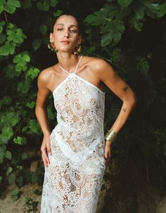 a woman wearing a white dress standing in front of some bushes and trees with her hands on her hips