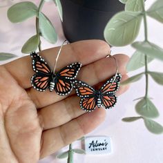 a hand holding two orange and black butterflies on it's earwires next to a potted plant