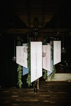 three white paper signs hanging from the ceiling in front of a black backdrop with greenery