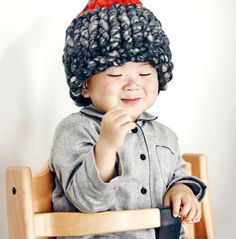 a little boy sitting in a wooden chair wearing a crocheted hat