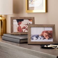 two framed pictures sitting on top of a wooden shelf next to a lamp and book