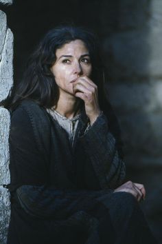 a woman sitting in front of a stone wall with her hand on her mouth and looking at the camera