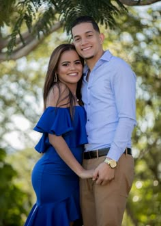 a man and woman posing for a photo under a tree in blue dress clothes with their arms around each other