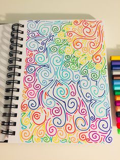 a spiral notebook with colored pencils next to it on a white surface and a drawing book in the background