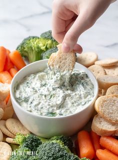 a hand dipping a cracker into a bowl of dip surrounded by veggies and crackers