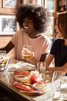 two women sitting at a table with food and wine in front of them, laughing