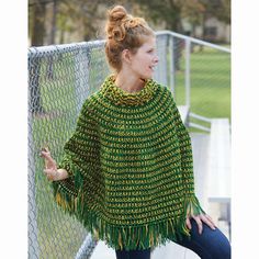 a woman standing next to a fence wearing a green knitted ponchy with fringes