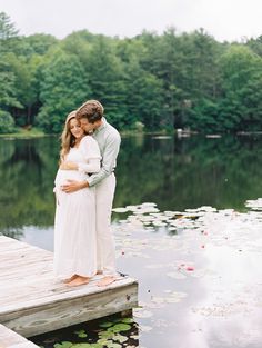 a pregnant couple standing on a dock in front of a lake with water lillies