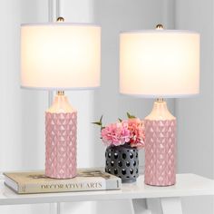 two pink lamps sitting on top of a white table next to a book and flowers