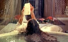 a woman laying in a hot tub reading a book with water pouring from the faucet behind her