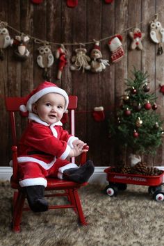 a baby in a santa suit sitting on a red chair next to a christmas tree