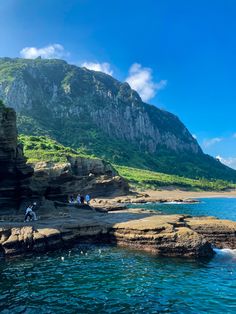 people are sitting on the rocks by the water and looking at the mountains in the distance