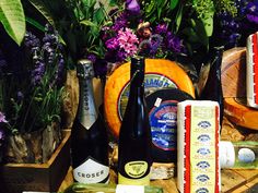 various cheeses and wine bottles on a table with purple flowers in the back ground