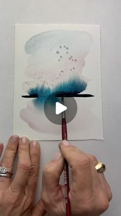 two hands holding a pencil and drawing on paper with watercolors in the background