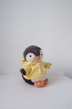 a stuffed penguin with a yellow shirt and scarf on it's head, sitting against a white background