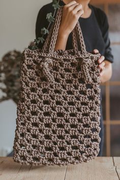 a woman holding a brown crocheted bag on top of a wooden table