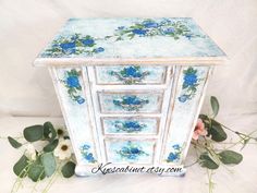 an old dresser with blue flowers painted on the top and bottom, sitting next to some green leaves