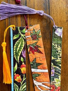 three colorful bookmarks with tassels hanging from them on a wooden table next to other books