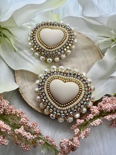 two white heart shaped earrings sitting on top of a piece of wood next to flowers