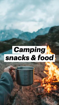 a person holding a cup in front of a campfire with mountains in the background