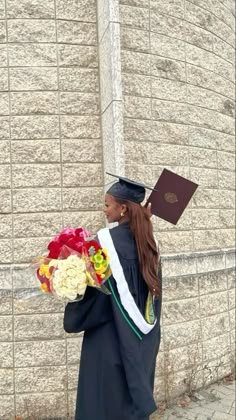 a woman in graduation gown holding flowers and a book standing next to a brick wall