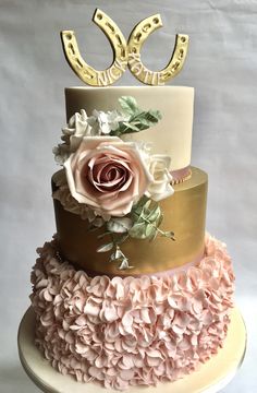 a three tiered cake with pink flowers and gold letters on top, sitting on a white pedestal