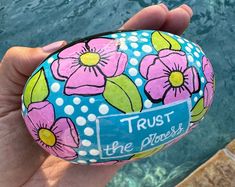 a hand holding a painted rock with flowers on it that says trust the shorer