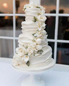 a white wedding cake with flowers on top is sitting in front of a large window