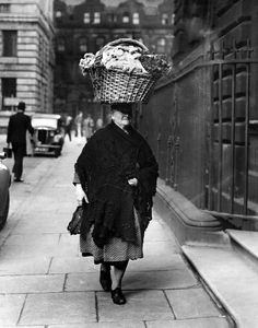 an old photo of a woman walking down the street with a basket on her head